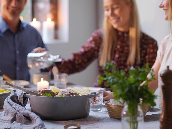 Tre personer sitter rundt et bord og spiser fårikål under et selskap, med grønne planter og lys i bakgrunnen.