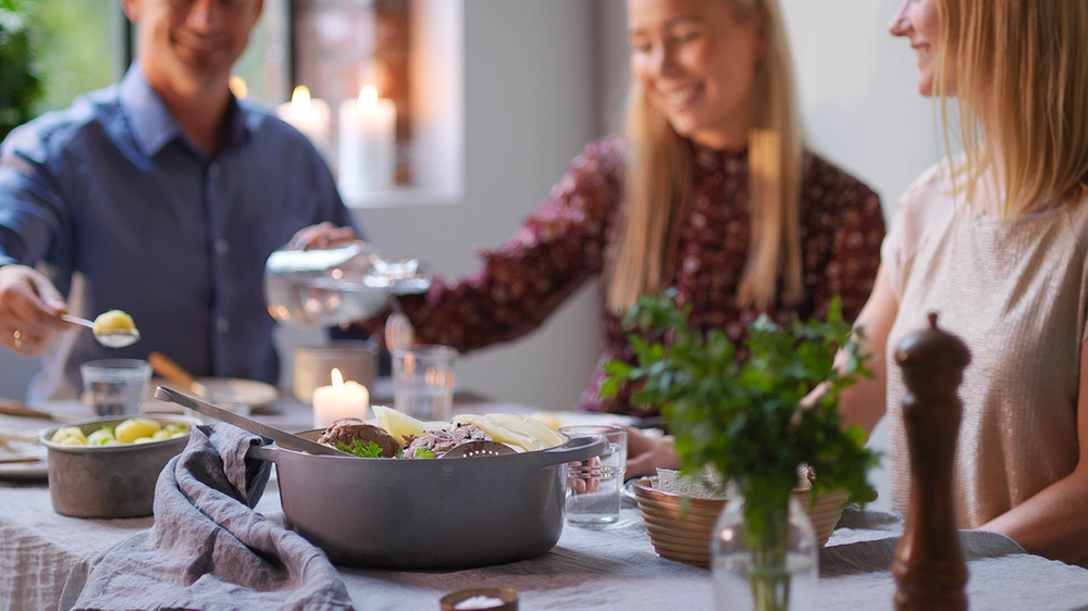 Tre personer sitter rundt et bord og spiser fårikål under et selskap, med grønne planter og lys i bakgrunnen.