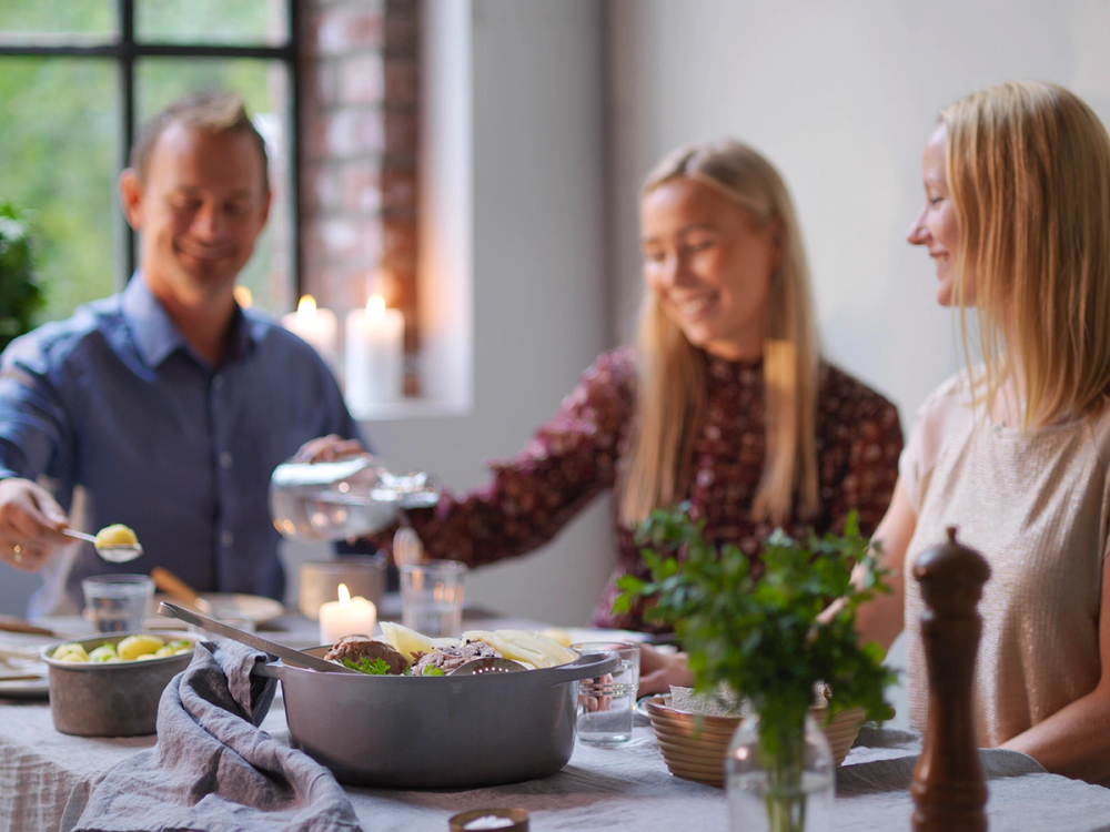 Tre personer sitter rundt et bord og spiser fårikål under et selskap, med grønne planter og lys i bakgrunnen.