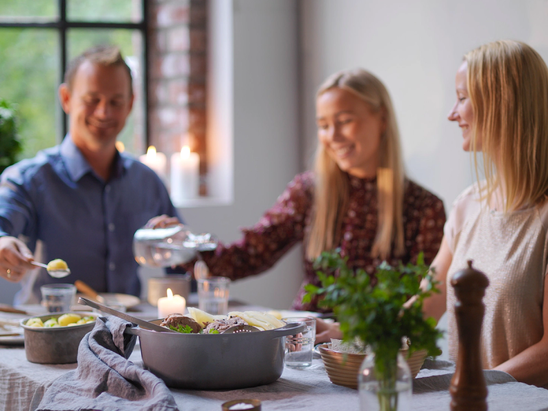 Tre personer sitter rundt et bord og spiser fårikål under et selskap, med grønne planter og lys i bakgrunnen.