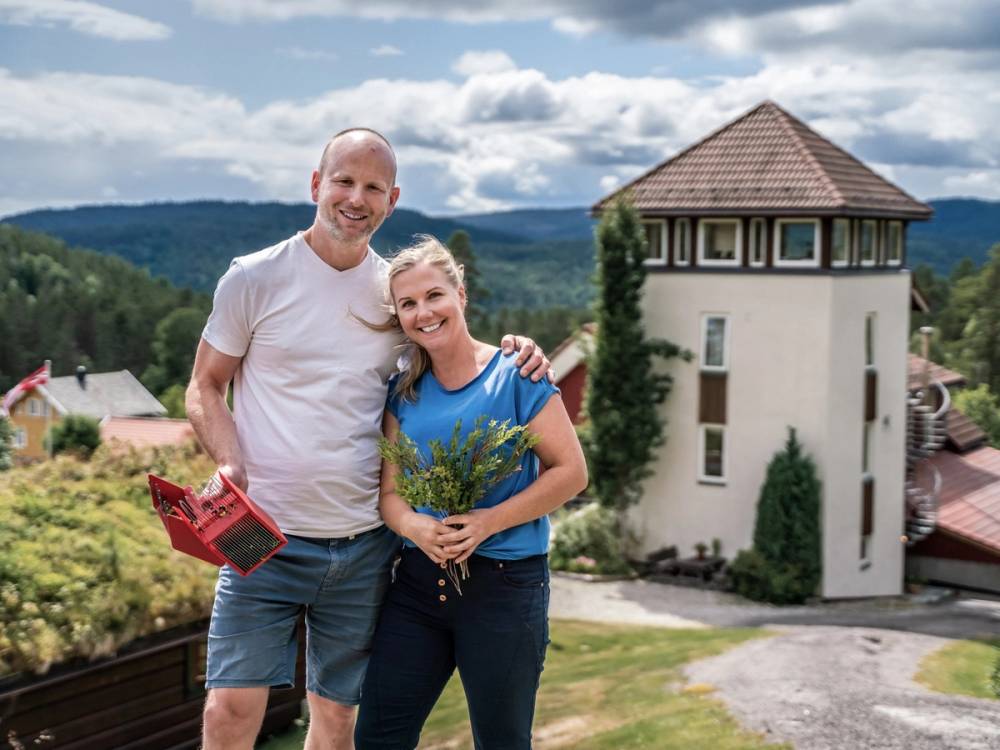 To personer står sammen foran et hus med en tårnformet del, omgitt av grønt landskap og fjell i bakgrunnen, med en skyet himmel.
