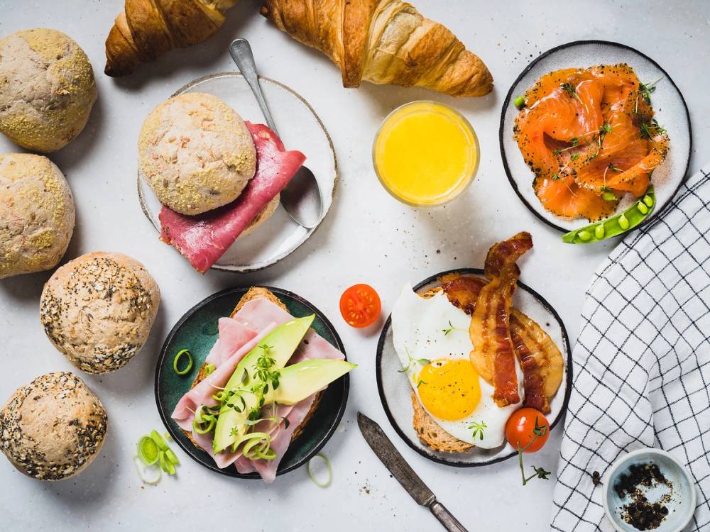 Frokostbord med brød, croissanter, pålegg, egg, grønnsaker og en kopp juice.