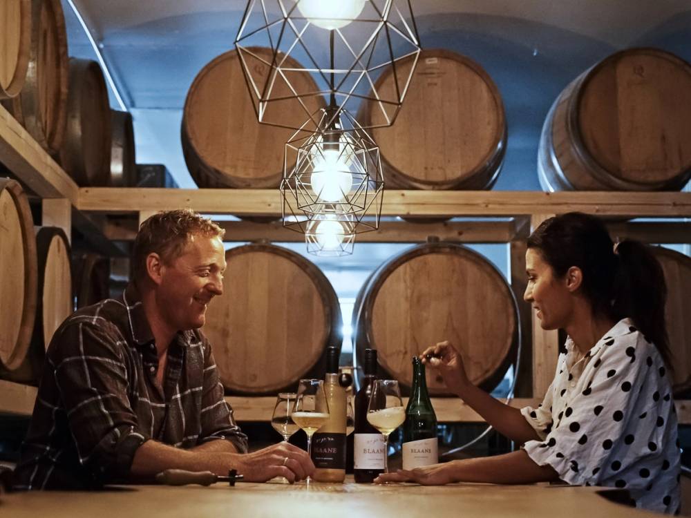 To personer som sitter og snakker over et bord med vinflasker i en vinkjeller med store tønner i bakgrunnen.