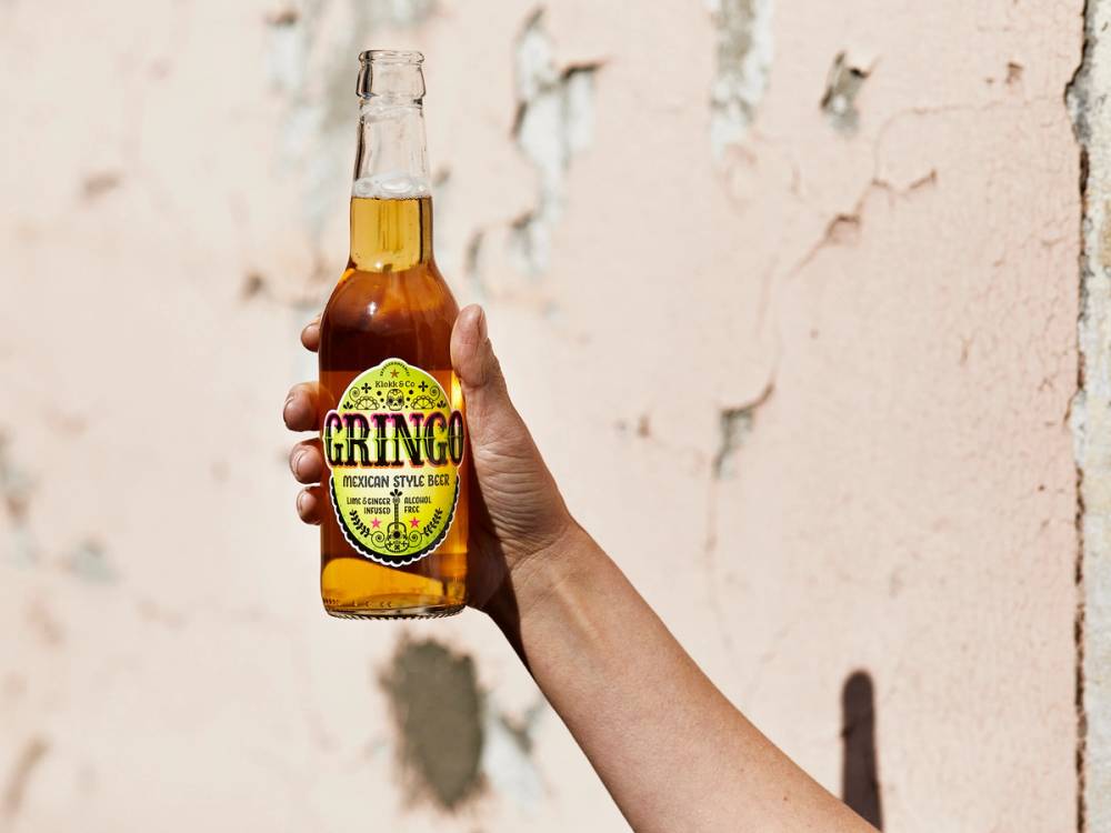 En person holder opp en flaske Gringo alkoholfritt øl foran en vegg med sprekkete maling.