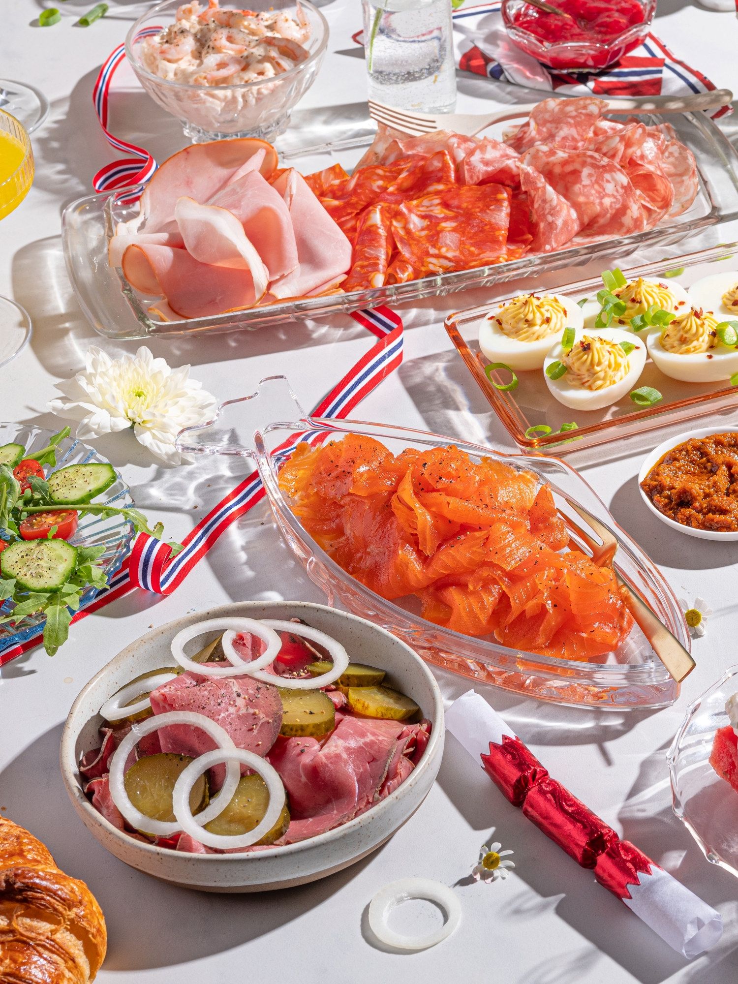 Frokostbord med ulike retter, inkludert spekemat, eggerøre, grønnsaker og pålegg, pyntet med 17. mai-sløyfer og blomster.