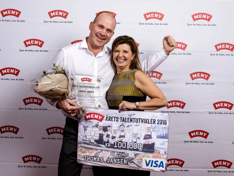 En person holder en stor sjekk og en annen person holder en pose, begge står foran en vegg med MENY-logoer, i forbindelse med Årets talentutvikler 2016.