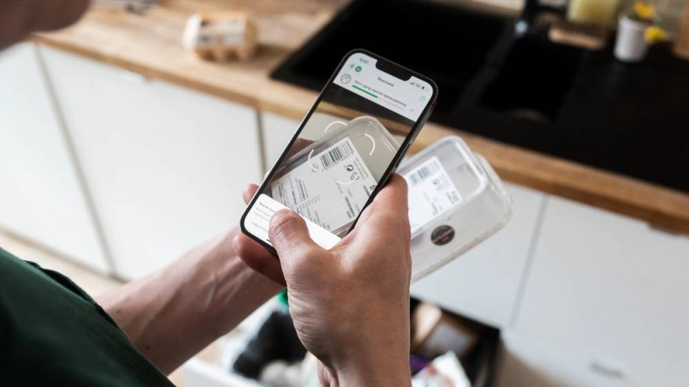 En person holder en smarttelefon over en kvittering på et kjøkkenbord, med en hvit kaffemaskin i bakgrunnen.