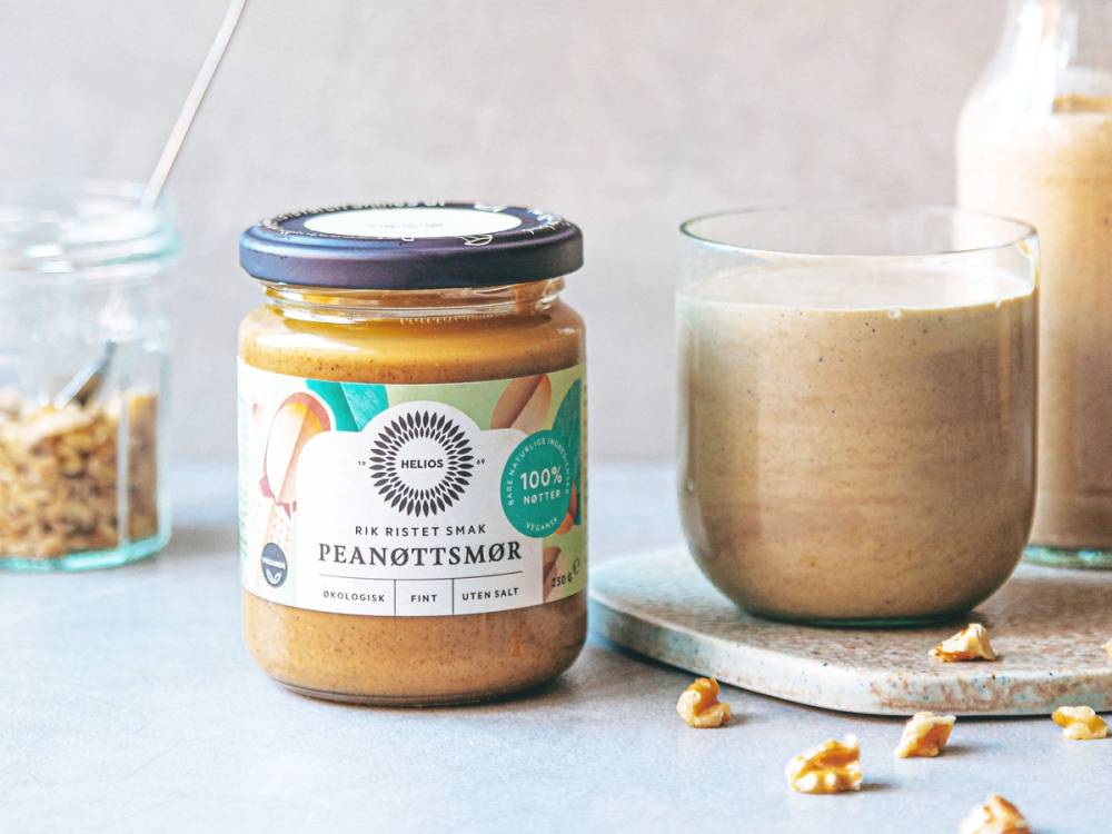 En glass med peanøttsmør smoothie og en glass med kaffe ved siden av på et kjøkkenbord, omgitt av nøtter og en glasskrukke med noe i bakgrunnen.