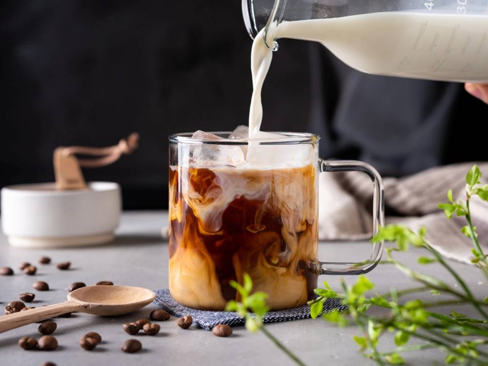 En iskald latte med melk som helles over kaffe i et klart glass, omgitt av kaffebønner og grønt bladverk på bordet.