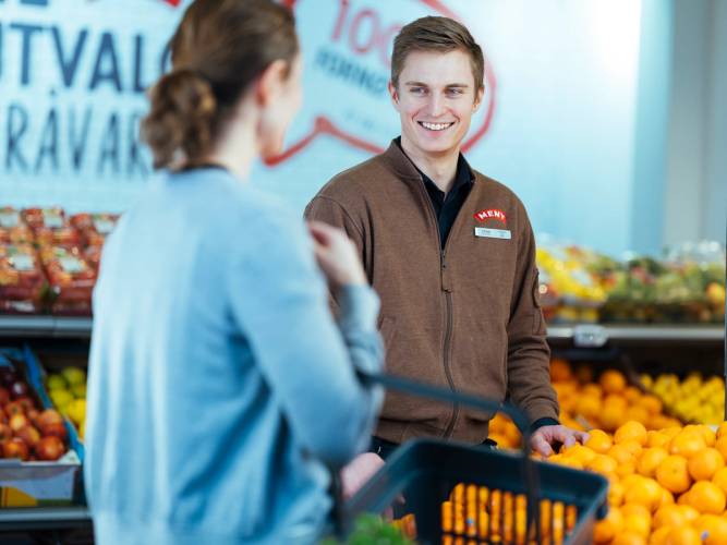 Kunde og ansatt i en dagligvarebutikk som snakker om sunnhet, med frukt og grønnsaker i bakgrunnen.
