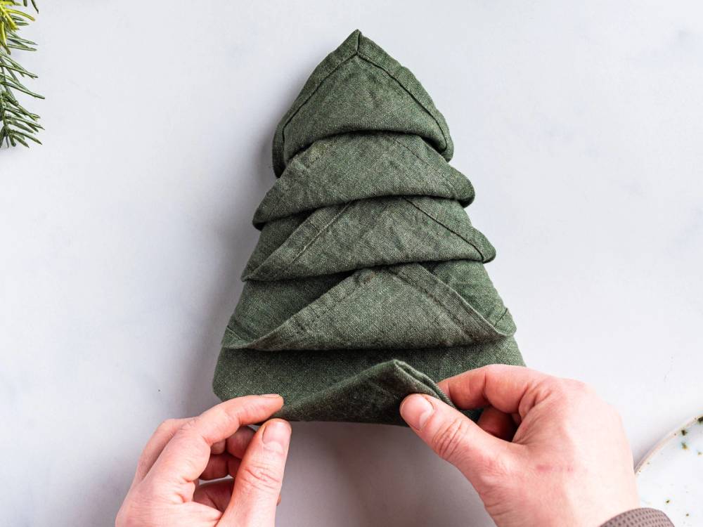 En person folder en grønn serviett i form av et juletre, med flere lag som ligner grener.