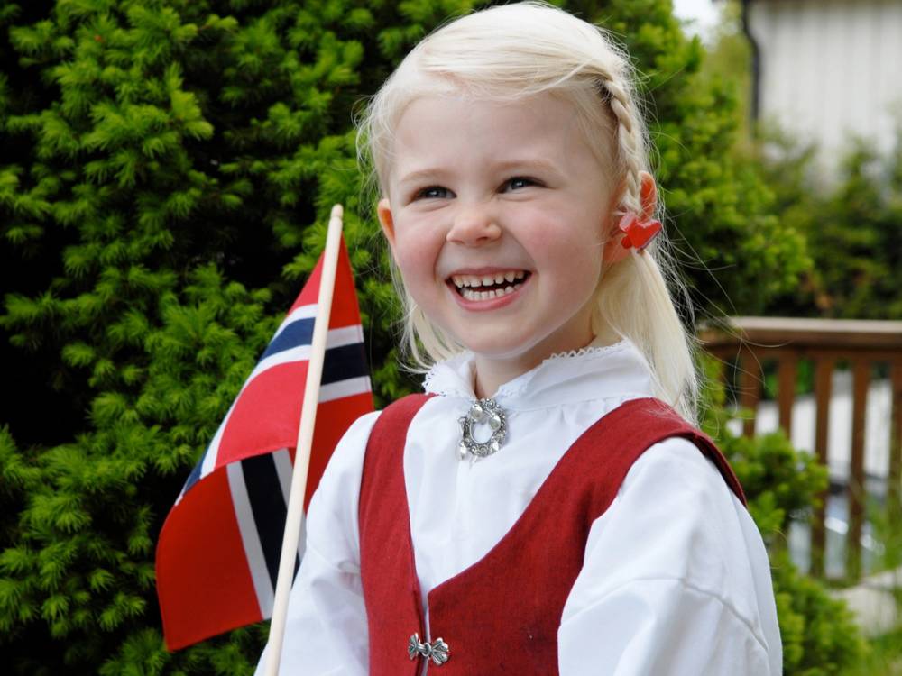 En person i tradisjonell bunad med en norsk flagg i hånden, ute i en hage eller park.