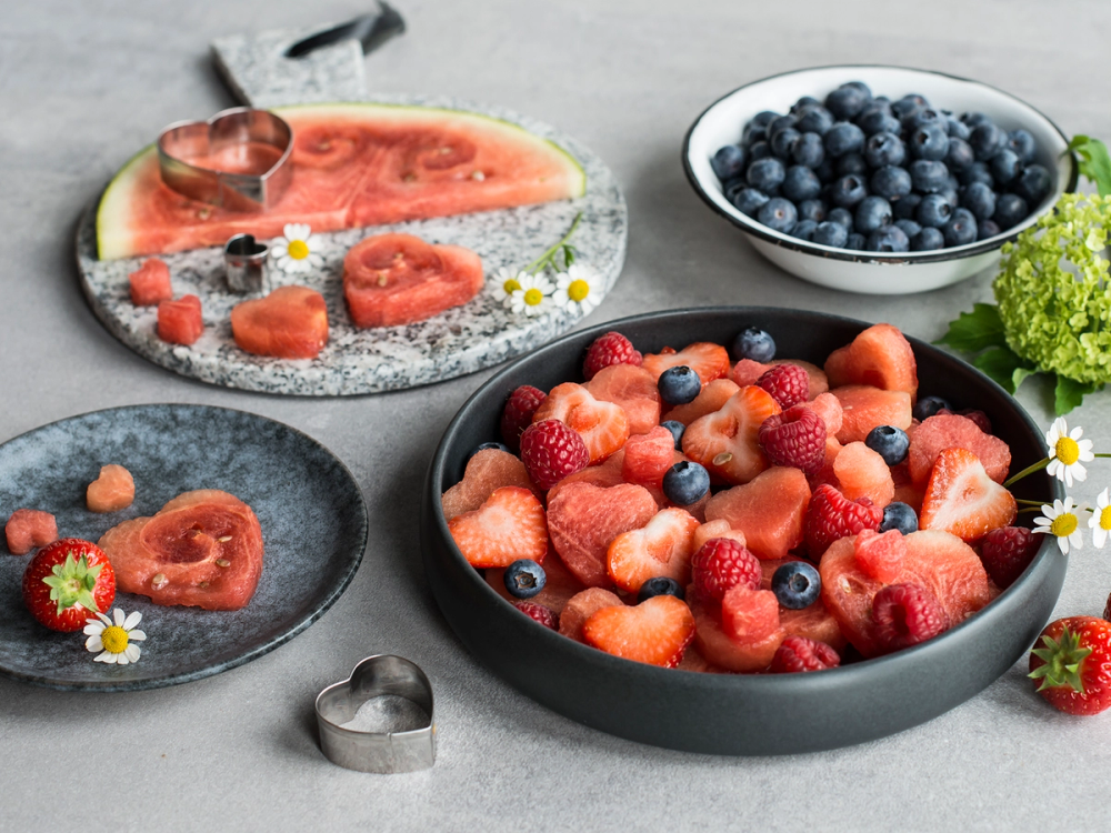 En skål med blandede bær, inkludert jordbær, blåbær og bringebær, sammen med en tallerken med vannmelonbiter og noen små blomster på et bord.