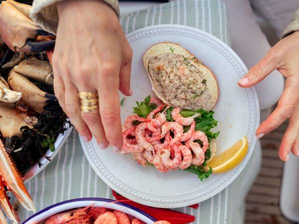 Hender som legger oppskåret reker og en skive brød med på en tallerken med sitronskive, på et bord dekket med et duk.