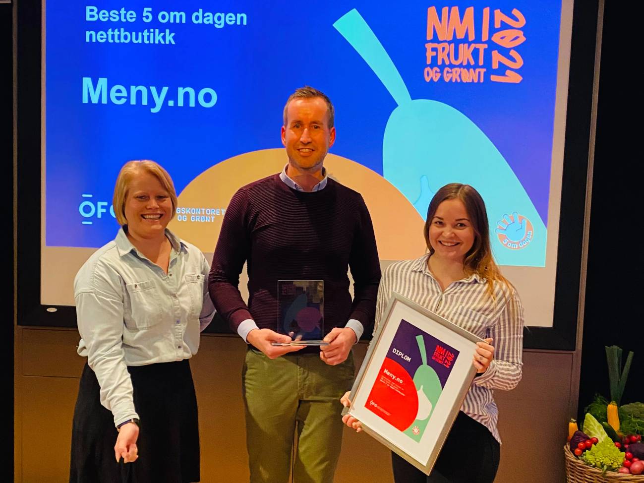 Tre personer står foran en skjerm som viser teksten 'Kåring NM 2021' og logoer, og de holder priser eller diplomer.