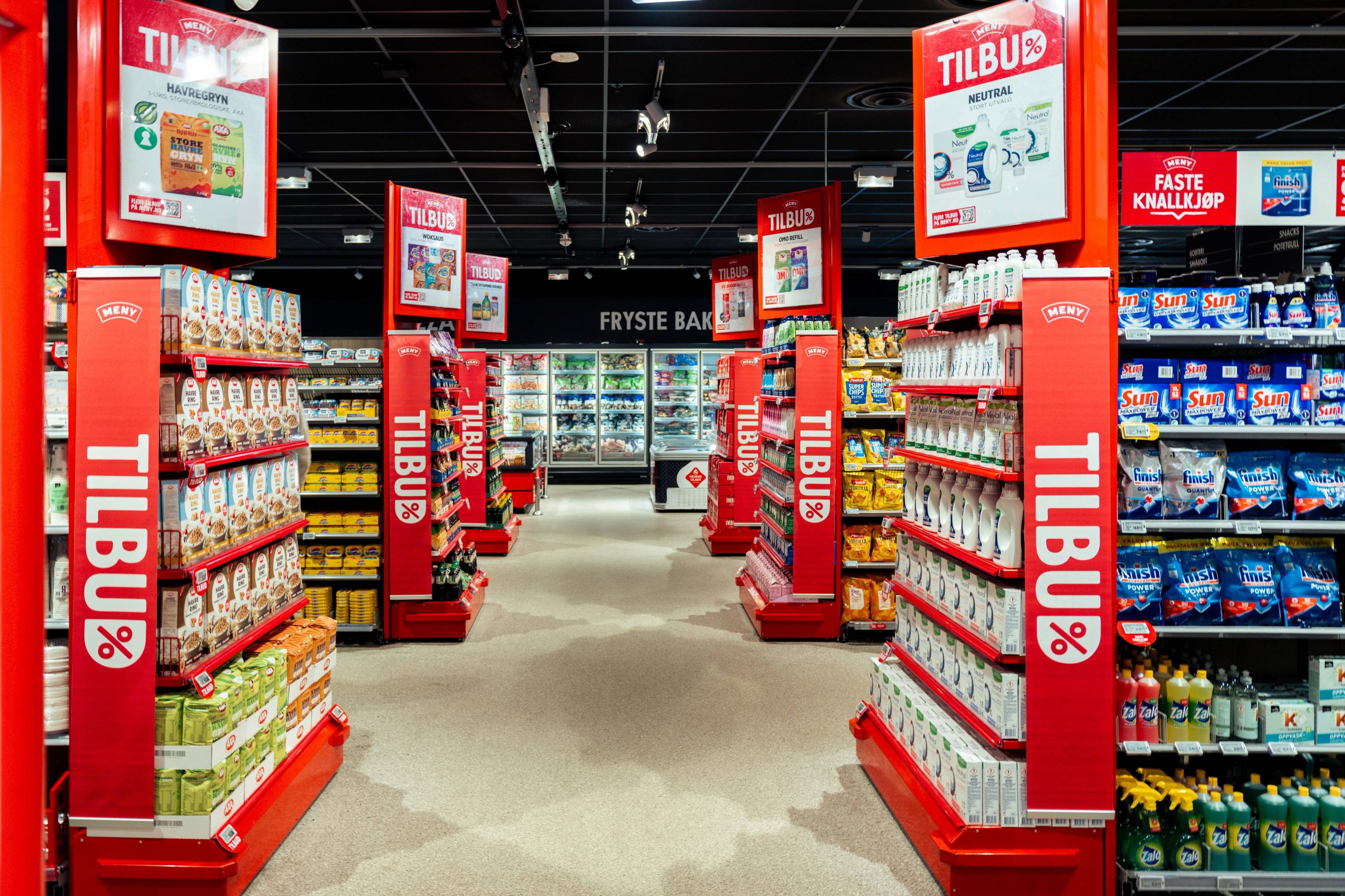 Inne i en dagligvarebutikk med hyller fylt med ulike produkter og tydelige TILBUD-skilt, sett fra midten av butikken mot kassen.