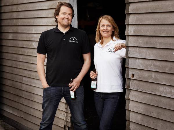 To personer står ved en trevegg, en av dem holder en flaske, og de har på seg klær med logoer.