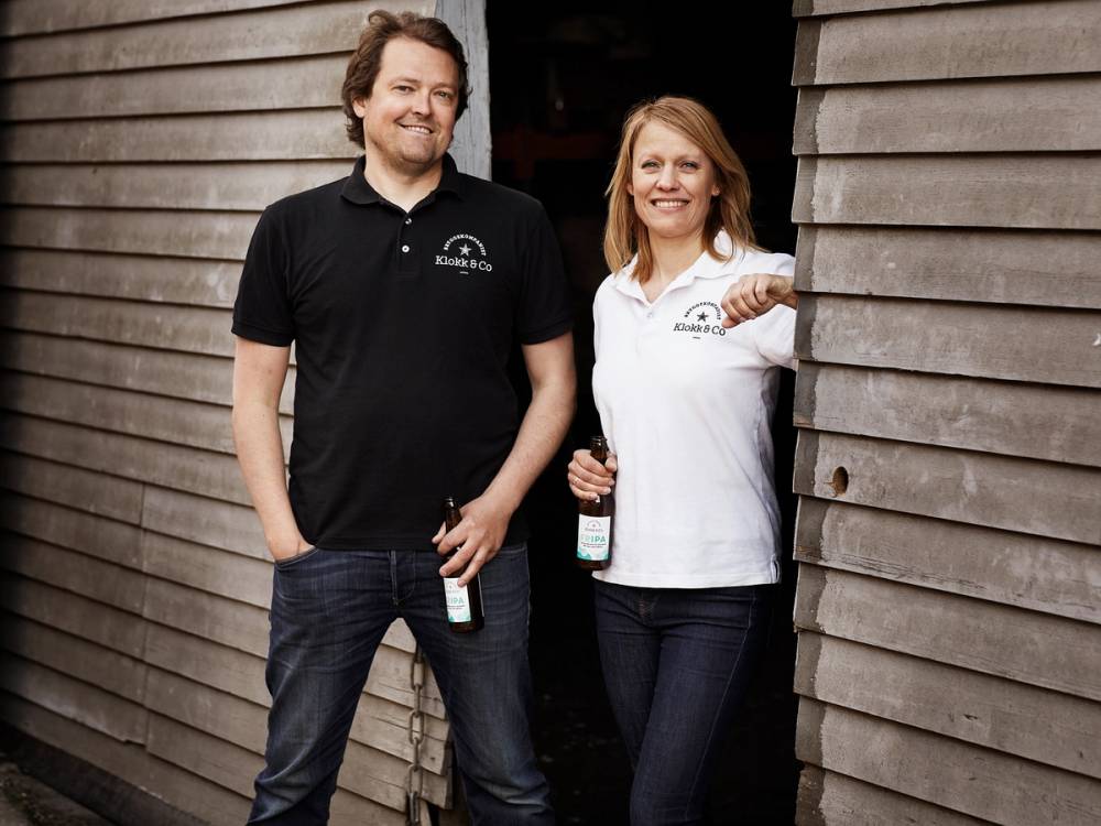 To personer står ved en trevegg, en av dem holder en flaske, og de har på seg klær med logoer.