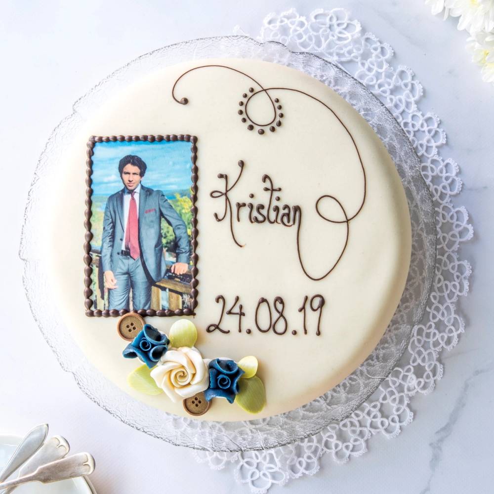 En konfirmasjonskake med bilde av en mann i dress, navn og dato skrevet på toppen, dekorert med små blomster og knapper, plassert på en hvit bordduk med blonder.