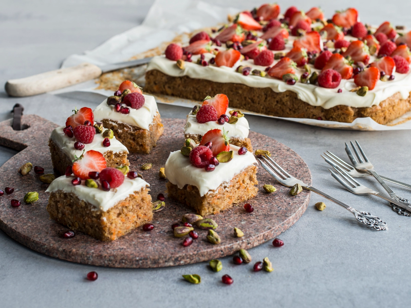 Gulrotkake i langpanne med krem og friske bær, skåret opp i firkanter på et fat med bestikk ved siden av.