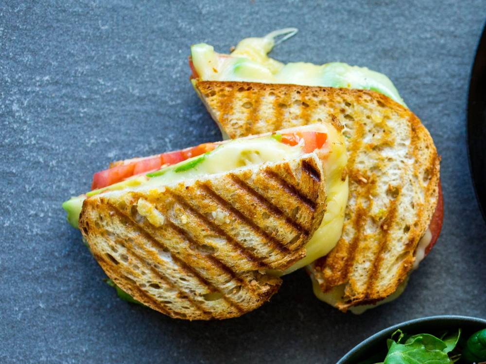 To ostesmørbrød med grillmerker, fylt med ost og skiver av tomat og agurk, på en mørk overflate.