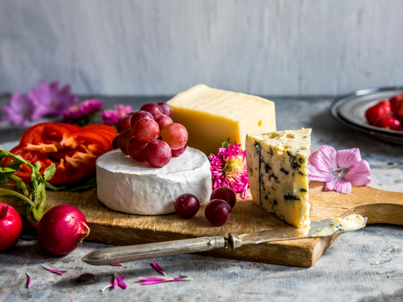 Ulike oster og druer på et trebrett med blomster rundt, foran en grå bakgrunn.
