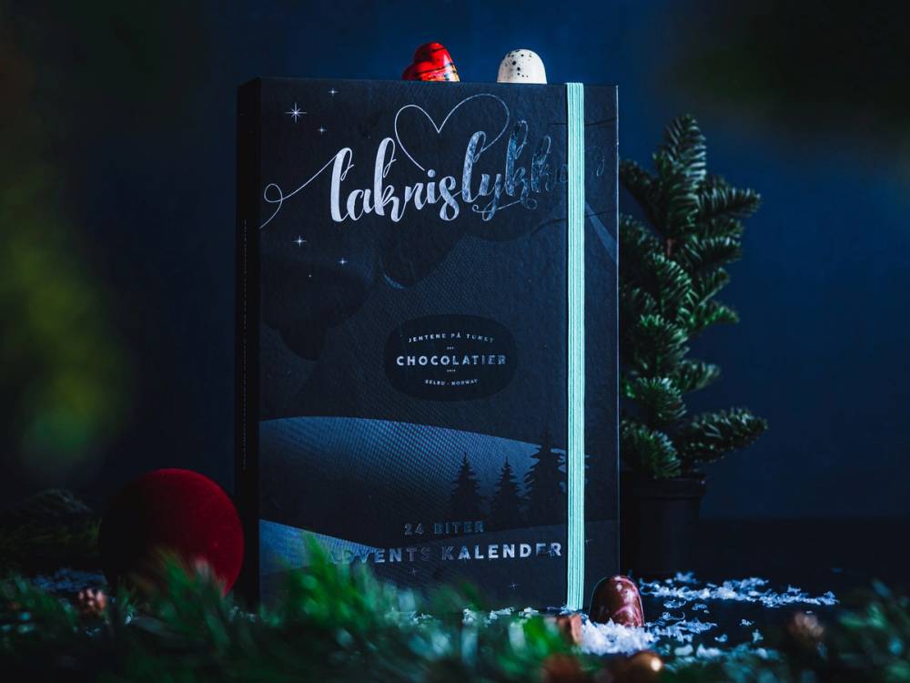 En kalender med tittelen 'Lakrids & Gløgg' plassert ved siden av en liten julegran og julepynt, med snø rundt.