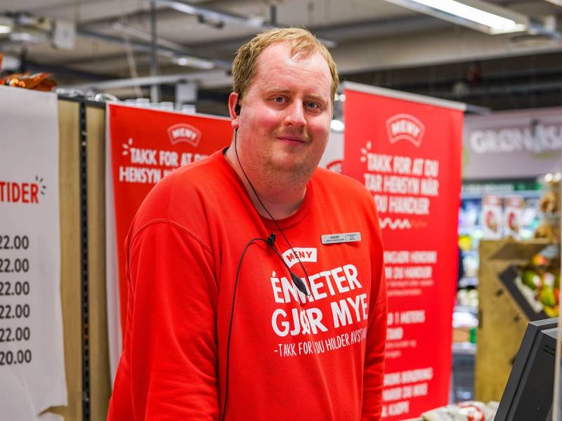 En person i rød uniform står i en butikk med reklameskilt i bakgrunnen, og holder en mikrofon eller lignende utstyr.