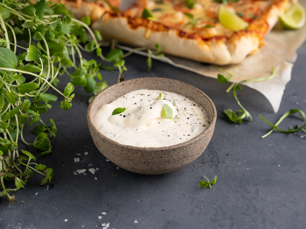 En skål med hvit pizzasaus omgitt av grønne urter og en pizza i bakgrunnen.