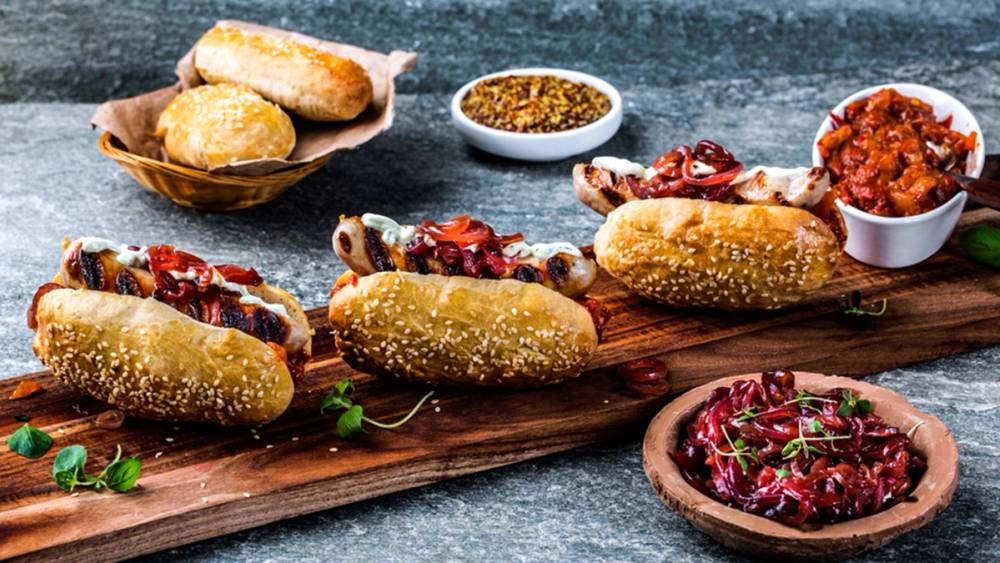 Fire pølser i brød med sennep og ketchup på et trebrett, sammen med små skåler med tilbehør og en bolle med rødkål.