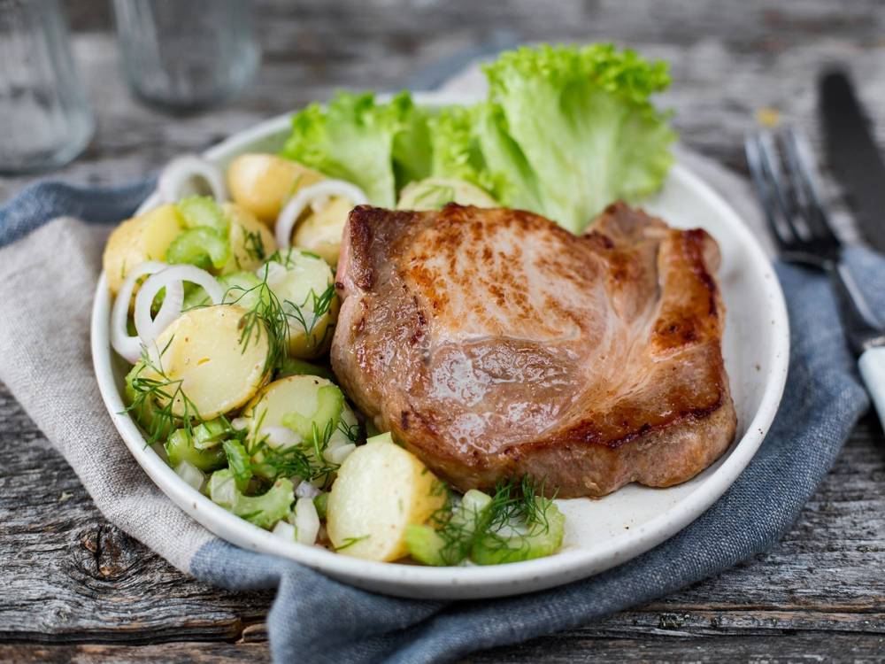 Svinekotelett med lun, fransk potetsalat og grønnsaker på en hvit tallerken.