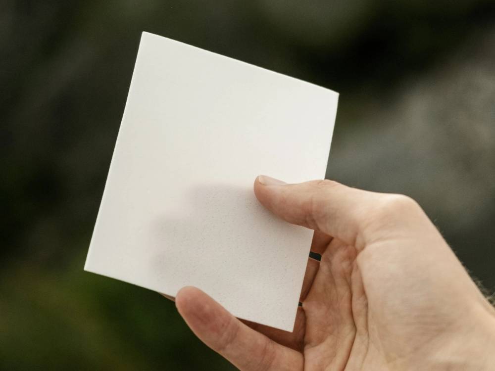 En person holder en tom, hvit kvadratisk lapp i hånden mot en utendørs bakgrunn.