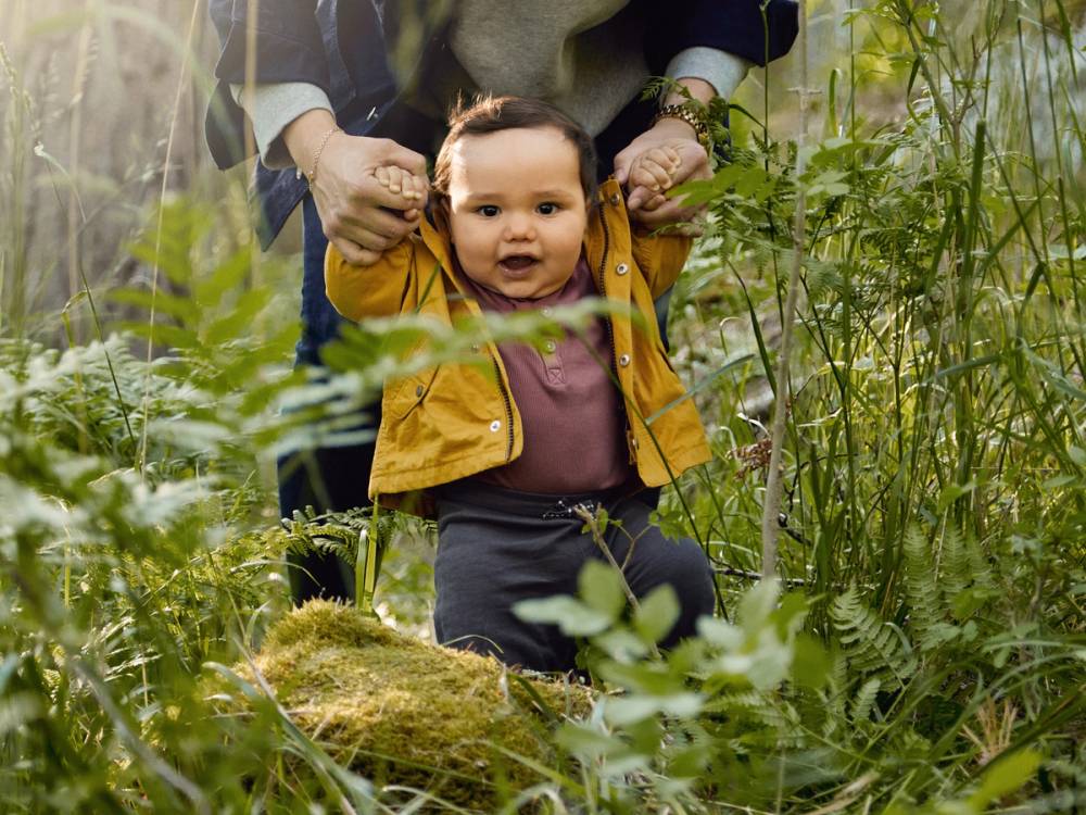 Et barn i gul jakke blir holdt av en voksen i en grønn skog eller park, med mye vegetasjon rundt.