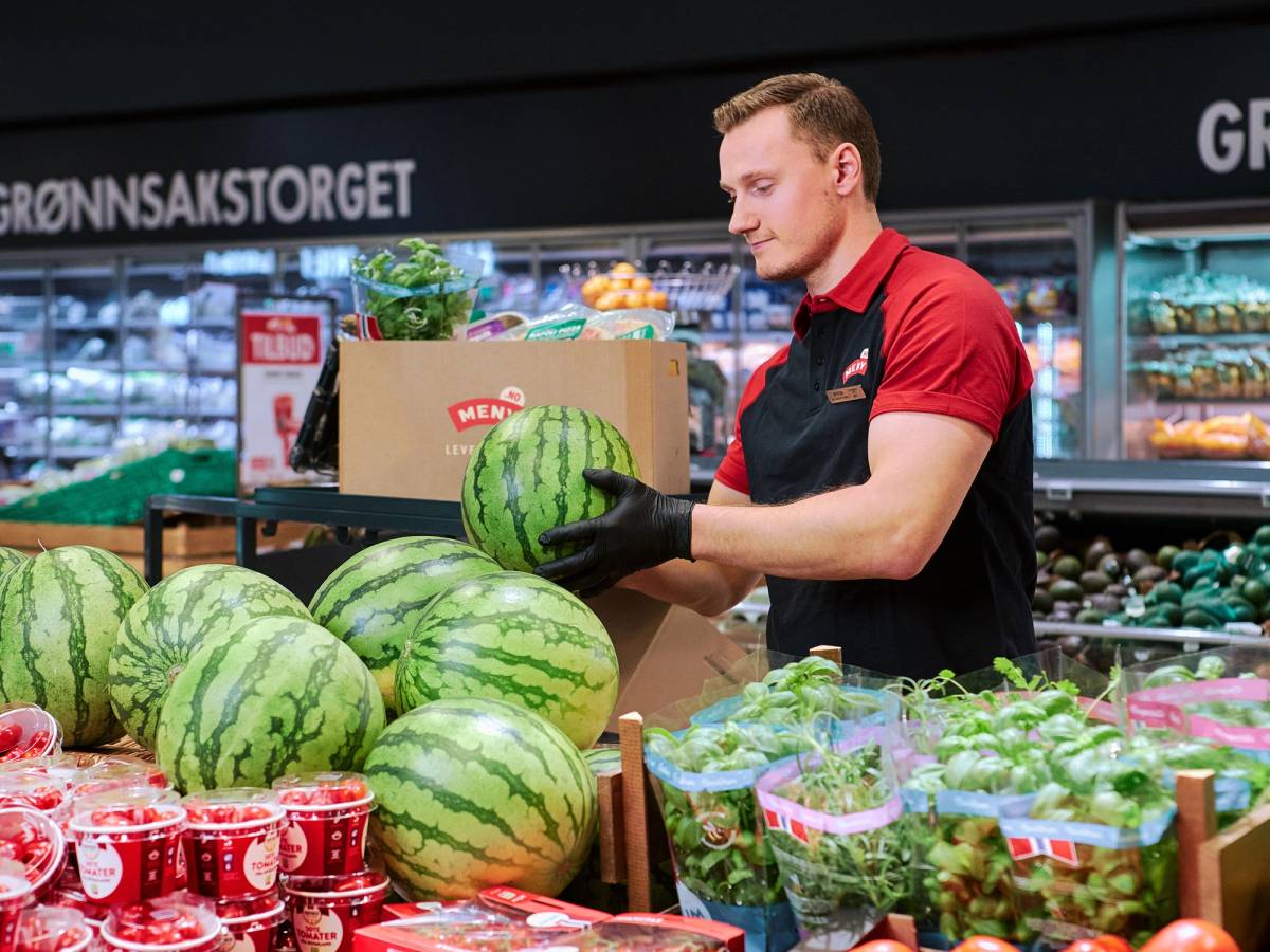 En mann i rød og svart uniform pakker vannmeloner i en butikk med grønnsaker og frukt i bakgrunnen.
