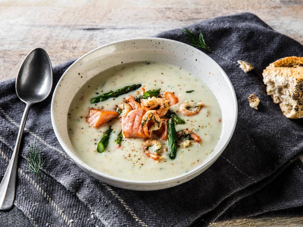 Løfotenfiskesuppe med grønnsaker og biter av fisk i en hvit skål på et bord med svart dukke, en skje ved siden av.