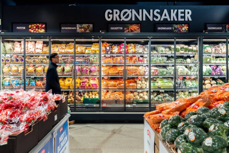 En person handler i grønnsaksavdelingen på MENY Tveita, med grønnsaker og frukt i kjøledisken og på bordet foran seg.