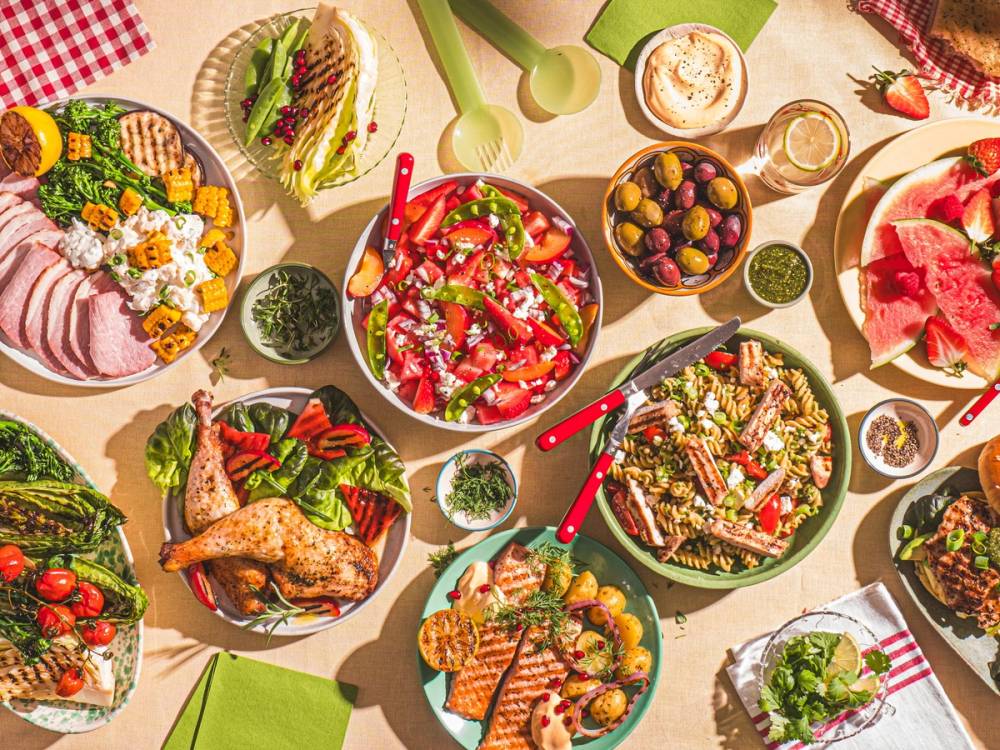 Et bord dekket med ulike sommerretter, salater og frukt, med grønne og røde farger, på en solrik dag.