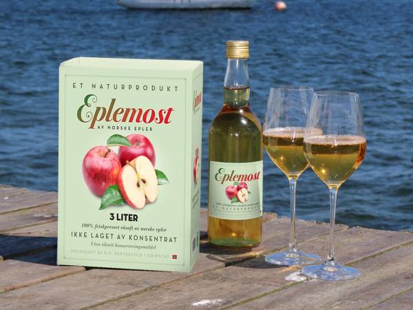 Eplemost og to glass med eplemost på en brygge ved vannet, med vann og båter i bakgrunnen.