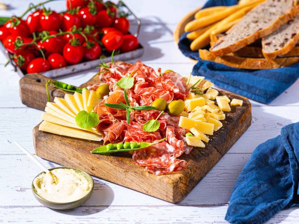 Et fat med ulike oster, skinke, grønne oliven og friske tomater på et trebrett, med brød og en liten skål med smør eller saus ved siden av.