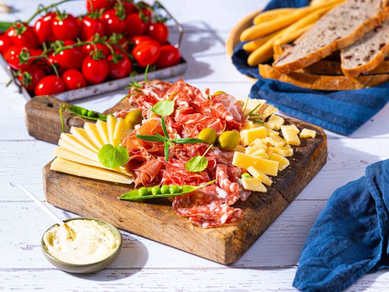 Et fat med ulike oster, skinke, grønne oliven og friske tomater på et trebrett, med brød og en liten skål med smør eller saus ved siden av.