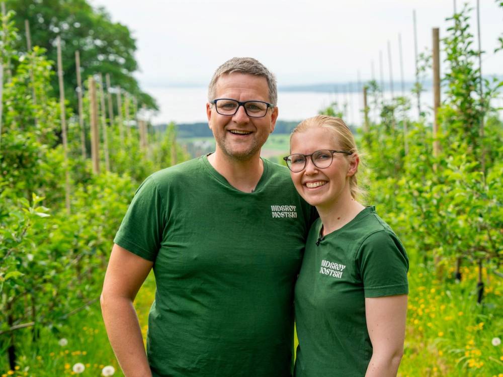 To personer i grønne T-skjorter står i en åker med grønne planter og trær i bakgrunnen.