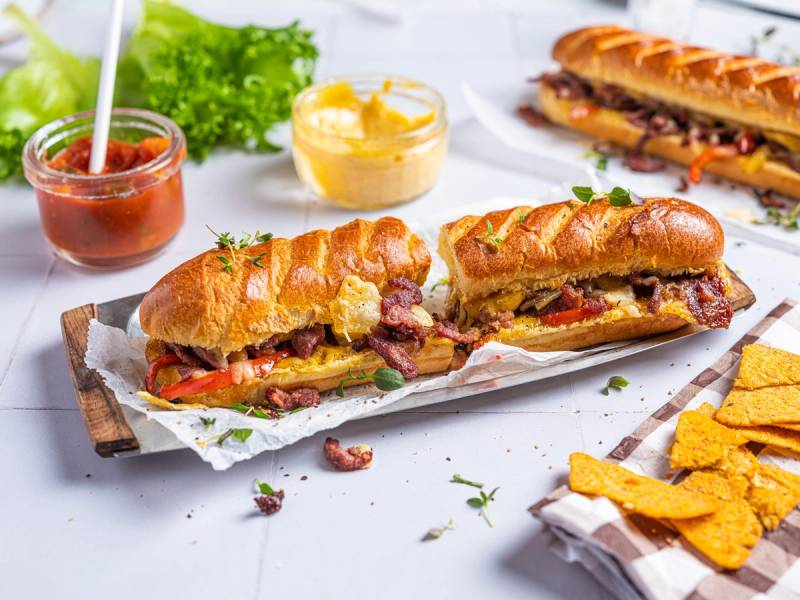 To biffsnadder sandwich med ost, bacon og grønnsaker, servert med chips og ulike sauser på en hvit bakgrunn.