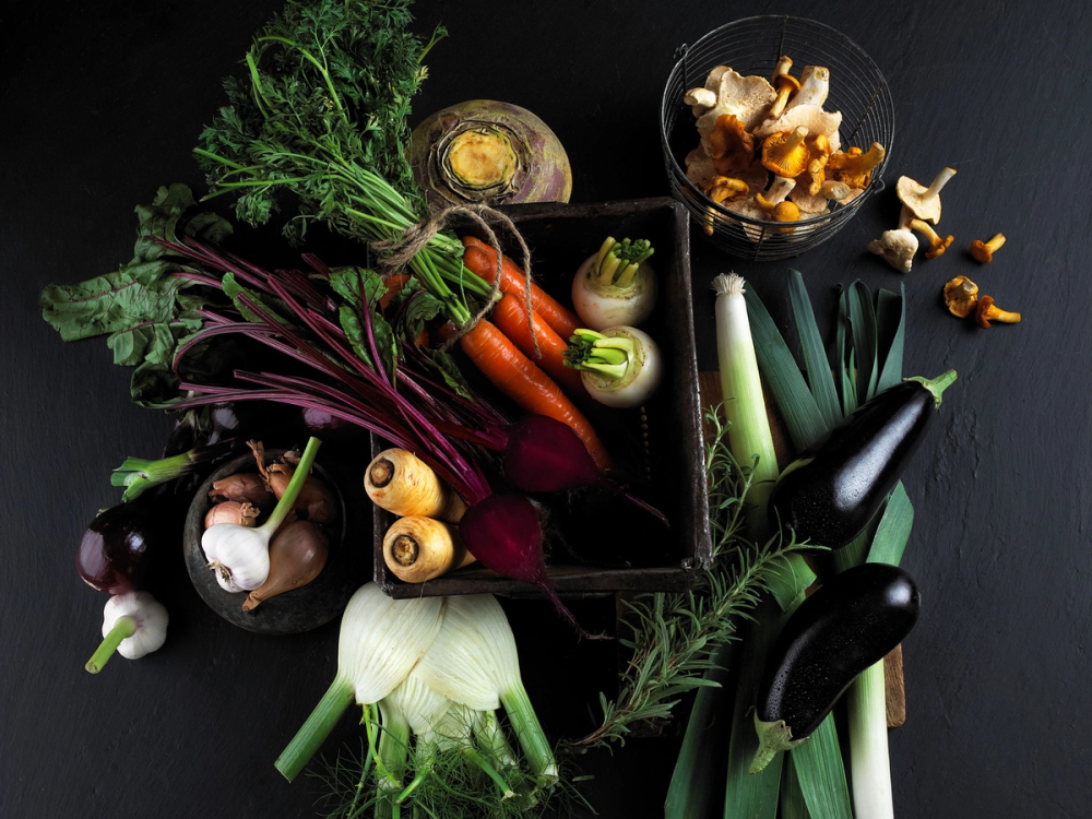 Et utvalg av ulike grønnsaker, inkludert gulrøtter, purre, sopp, kål og eggplanter, lagt ut på en svart bakgrunn.