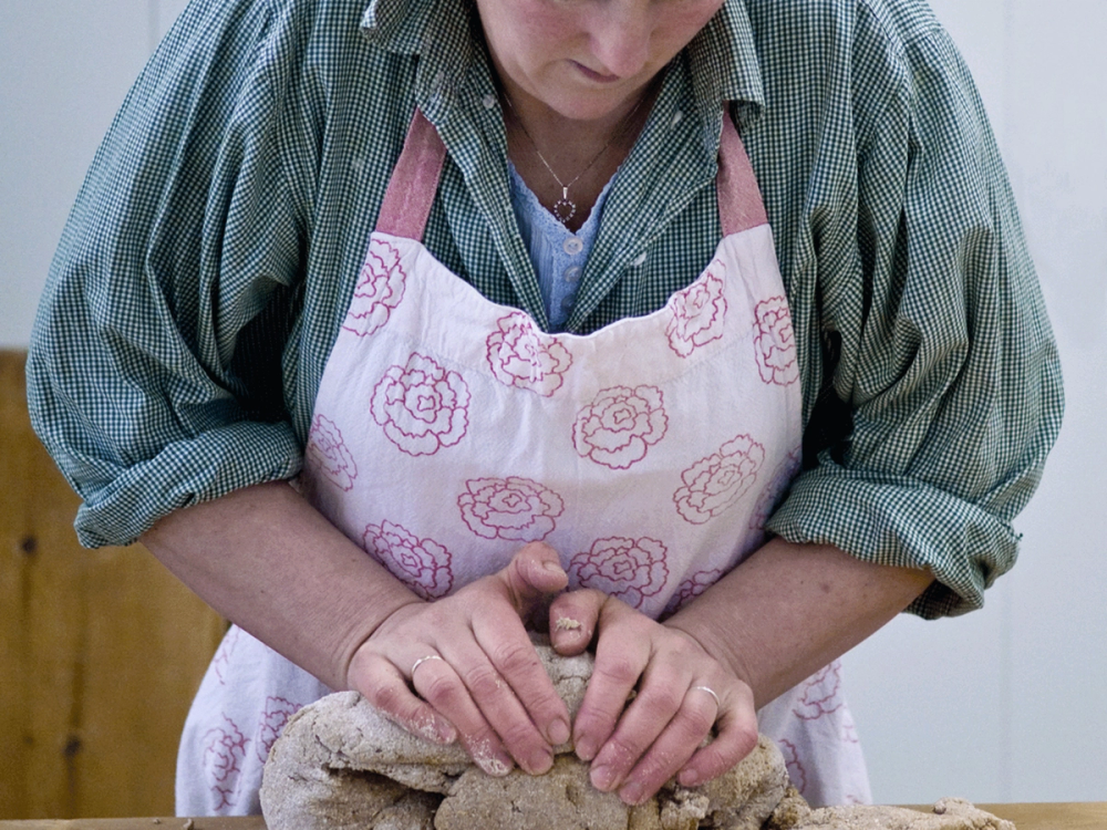 En person baker med fokus på hendene som former deigen, iført en rosa og hvit forklær med blomsterdesign.
