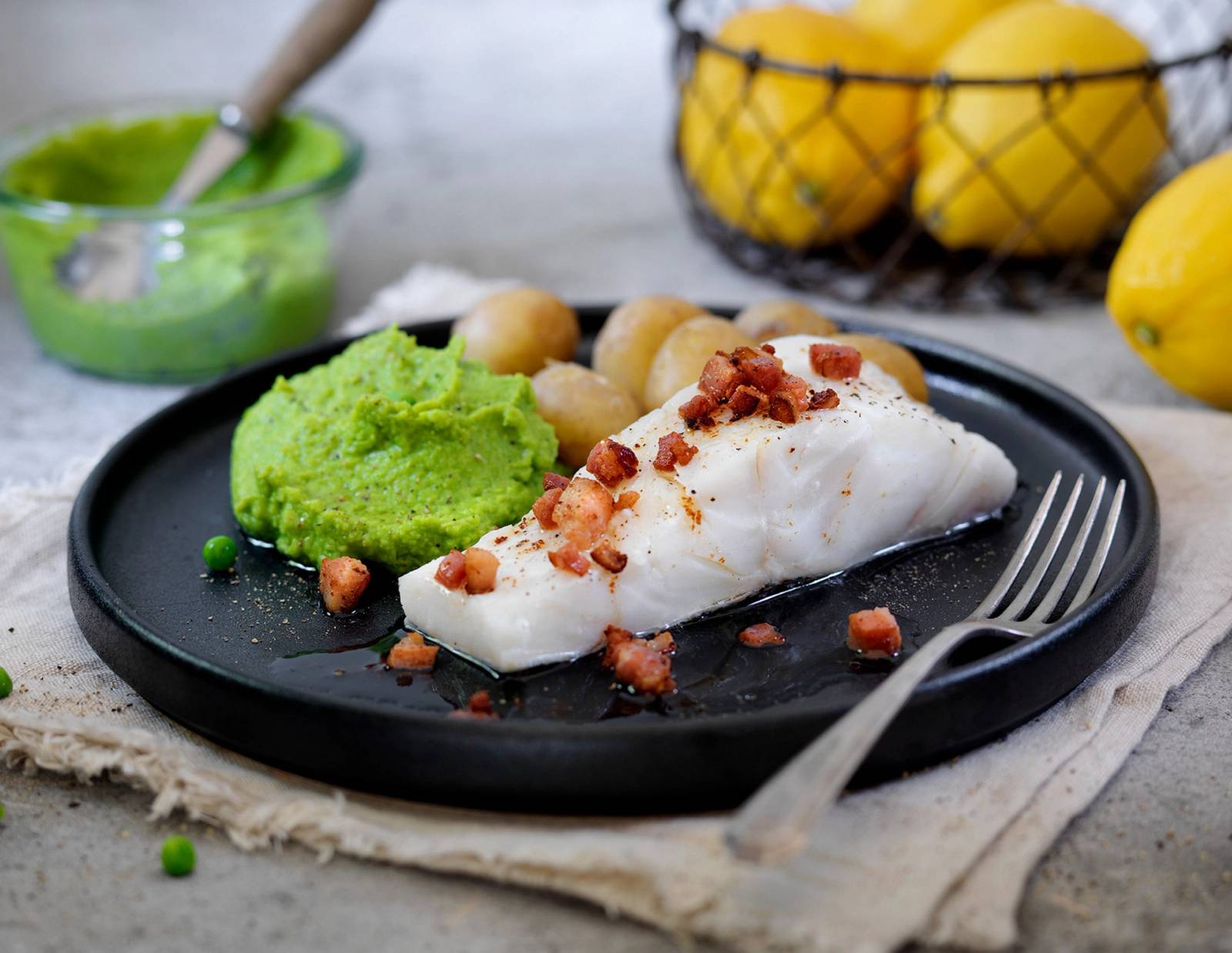 Lettsaltet torsk med ertepure og bacon på en svart tallerken, med en liten skål med grønn ertepure ved siden av.