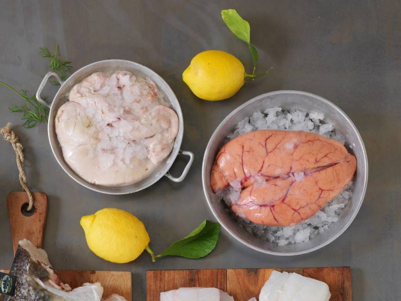 Skrei med rogn på en tallerken, omgitt av sitroner og urter, på et kjøkkenbord.