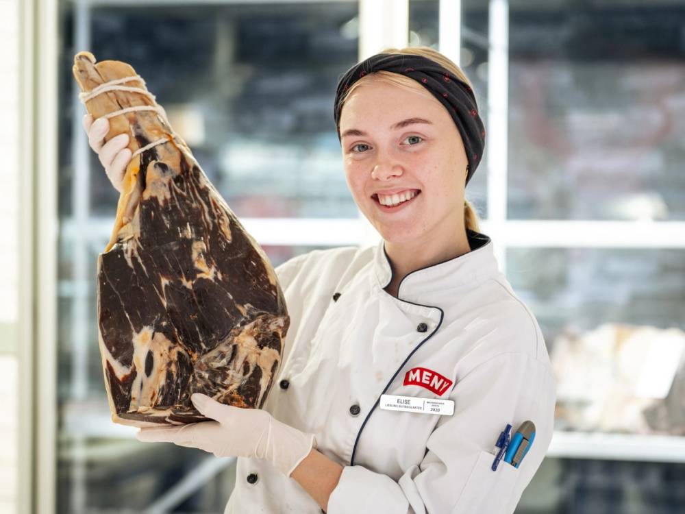 En person i hvit kjøkkenskjorte holder opp en stor skinke eller kjøttstykke i en butikk eller kjøkkenmiljø.