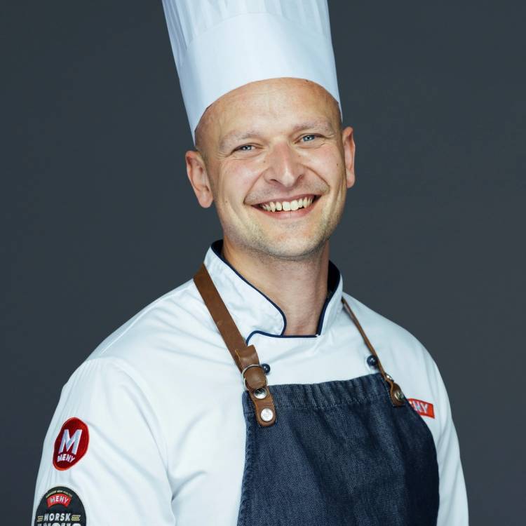 En kok i hvit kokkeuniform og blå forkle står mot en mørk bakgrunn, med ansiktet dekket av en beige firkant.