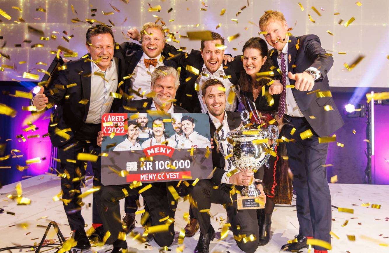 En gruppe personer feirer med pokaler og en sjekk etter å ha vunnet en pris, med gullkonfetti i luften.