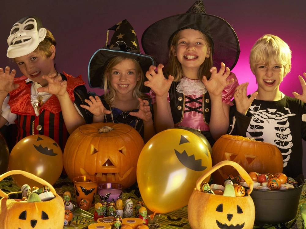 Barn i Halloween-kostymer med blodig popcorn og pyntede gresskar foran en rosa bakgrunn.